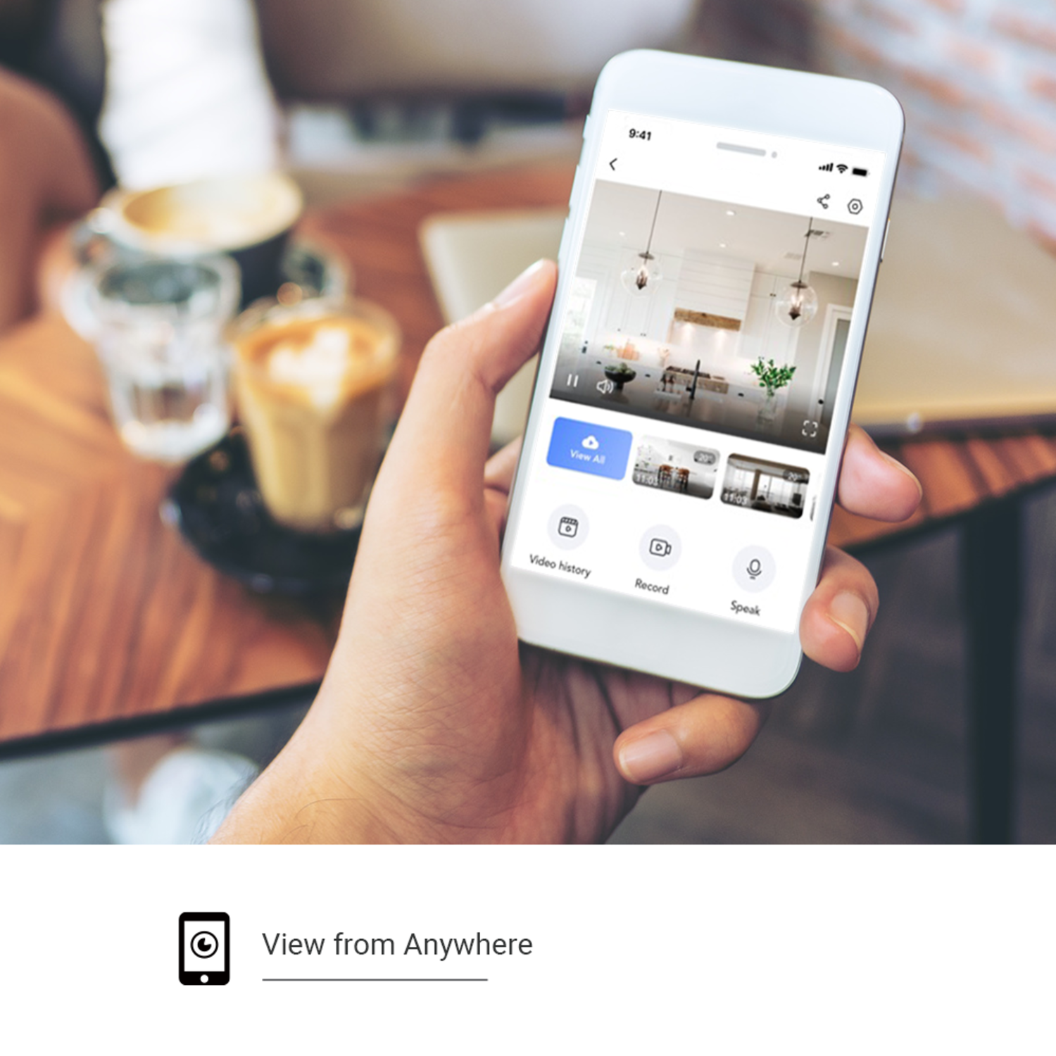 A hand holding a phone displaying a kitchen view. Coffee cups are in the background. The phone shows a security app.