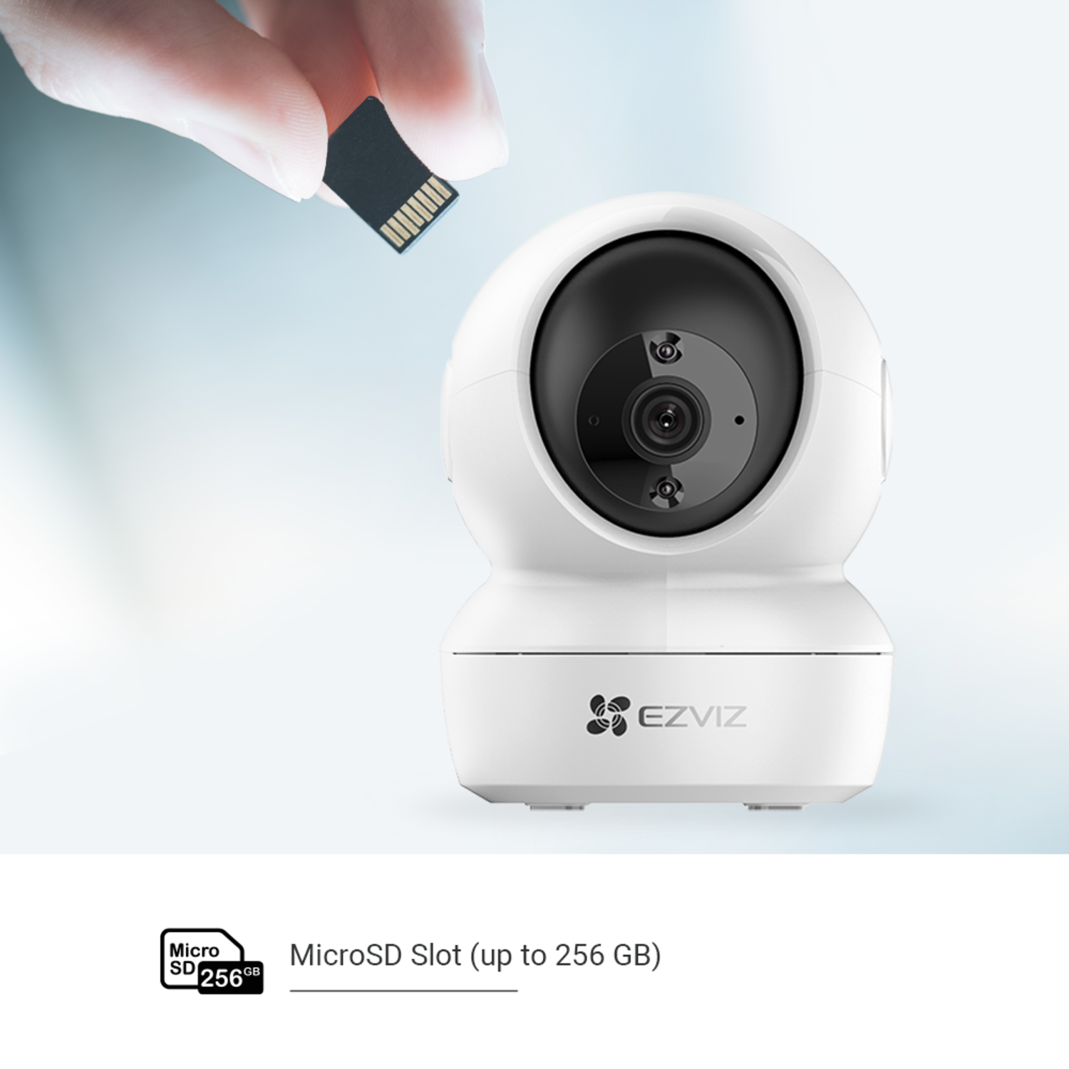 A hand holding an SD card above a white EZVIZ security camera. The camera has a black lens and is on a light blue background.