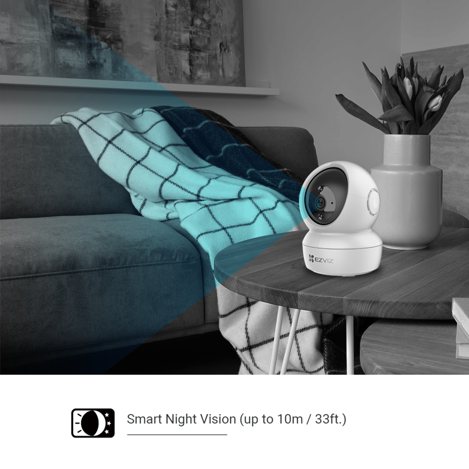 A white security camera sits on a table. A vase of flowers is next to it. A blanket is draped over a sofa in the background.