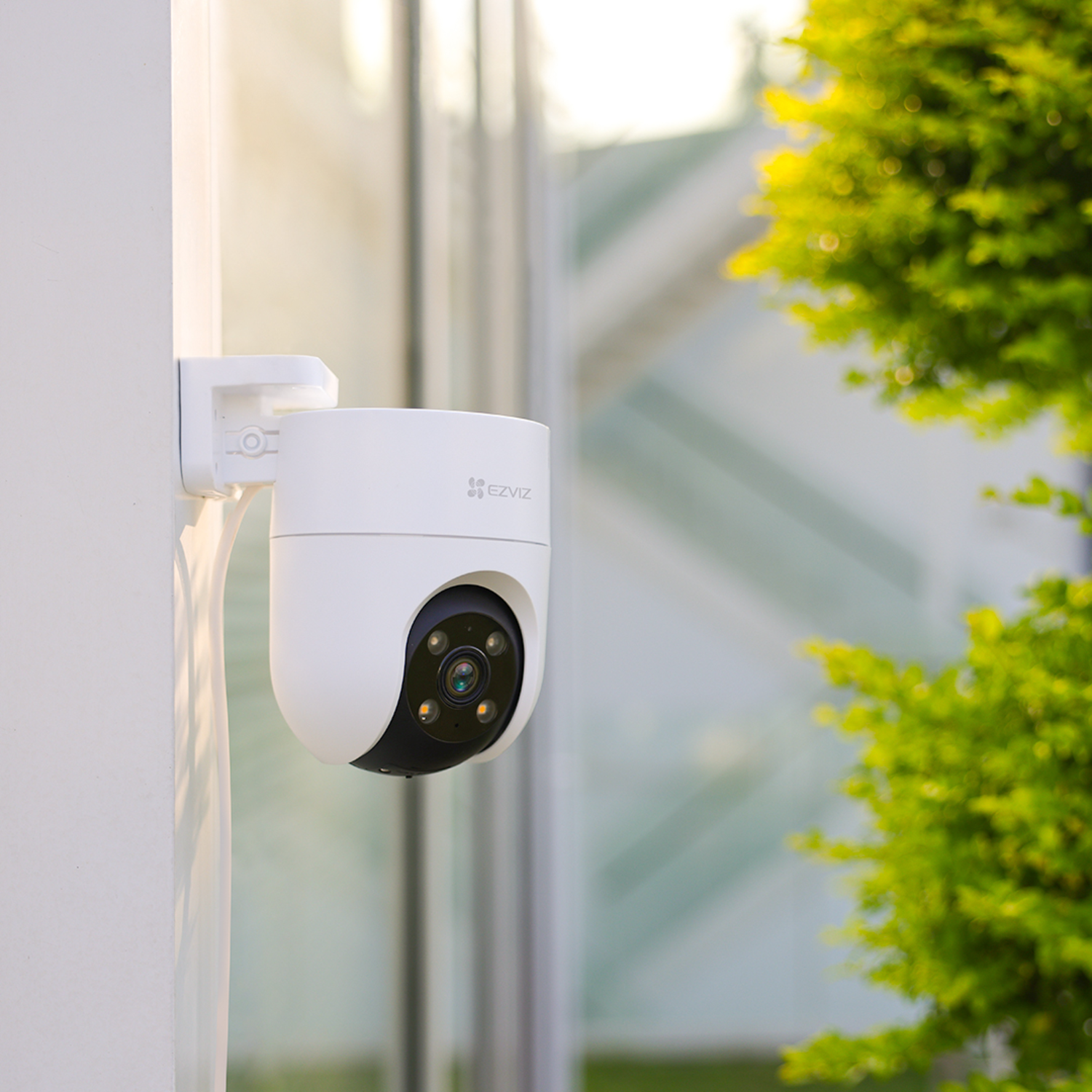 A white security camera is mounted on a white wall. Green foliage is in the background.