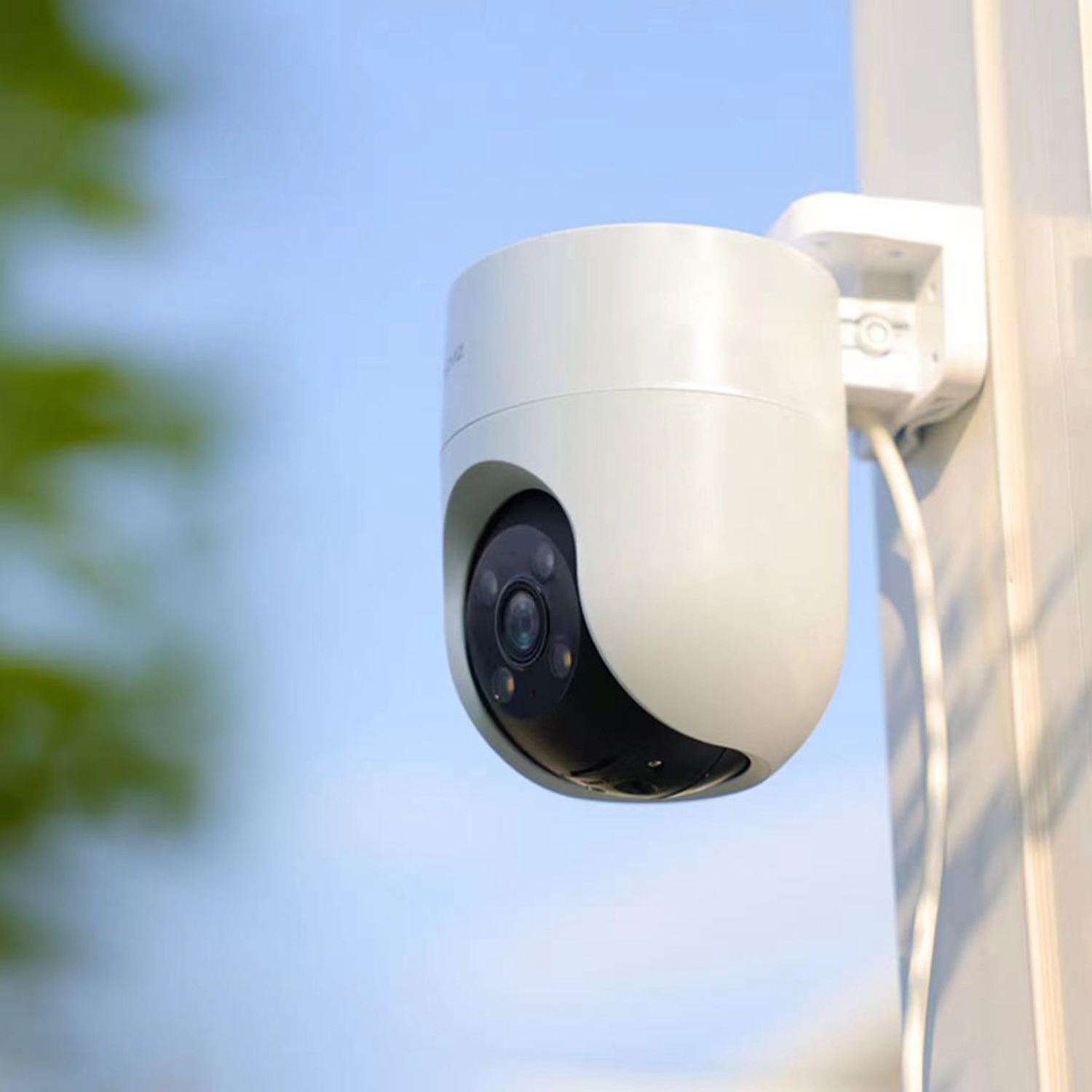 A white security camera mounted on a pole against a blue sky. The camera has a dark lens and is angled downwards.