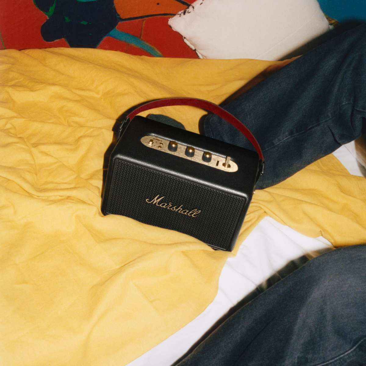 Een zwarte Marshall-luidspreker met een rode handgreep ligt op een geel bed. Jeans en een kleurrijke deken op de achtergrond.