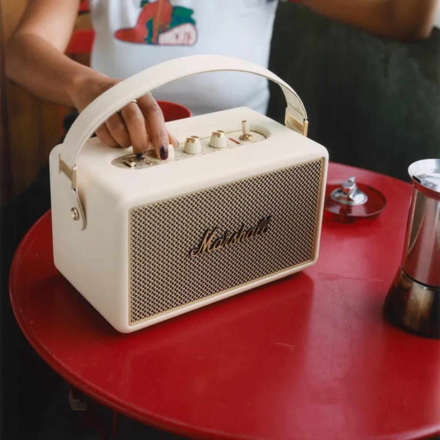 Een persoon past een witte Marshall-luidspreker aan op een rode tafel. De luidspreker heeft een handvat en gouden accenten. Er staat een koffiezetapparaat in de buurt.
