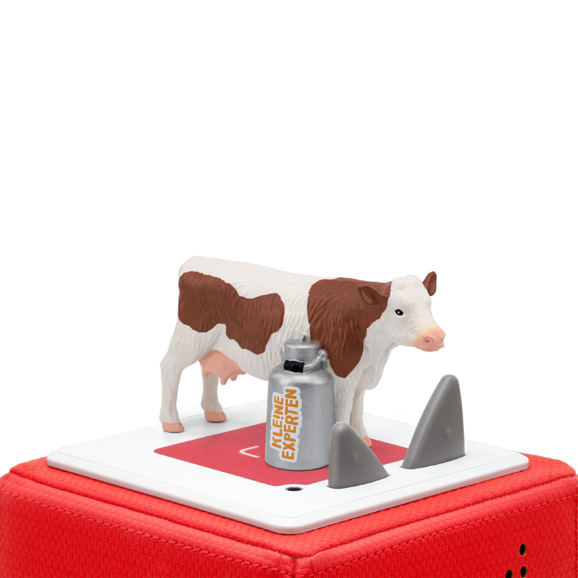 Ein Spielzeugkuh mit braunen Flecken steht auf einer weißen Oberfläche. Milchkännchen und graue Formen sind in der Nähe.