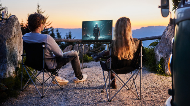 Zwei Personen schauen draußen auf einen Bildschirm. Im Hintergrund sind ein Van und Berge zu sehen. Der Bildschirm zeigt einen Astronauten.