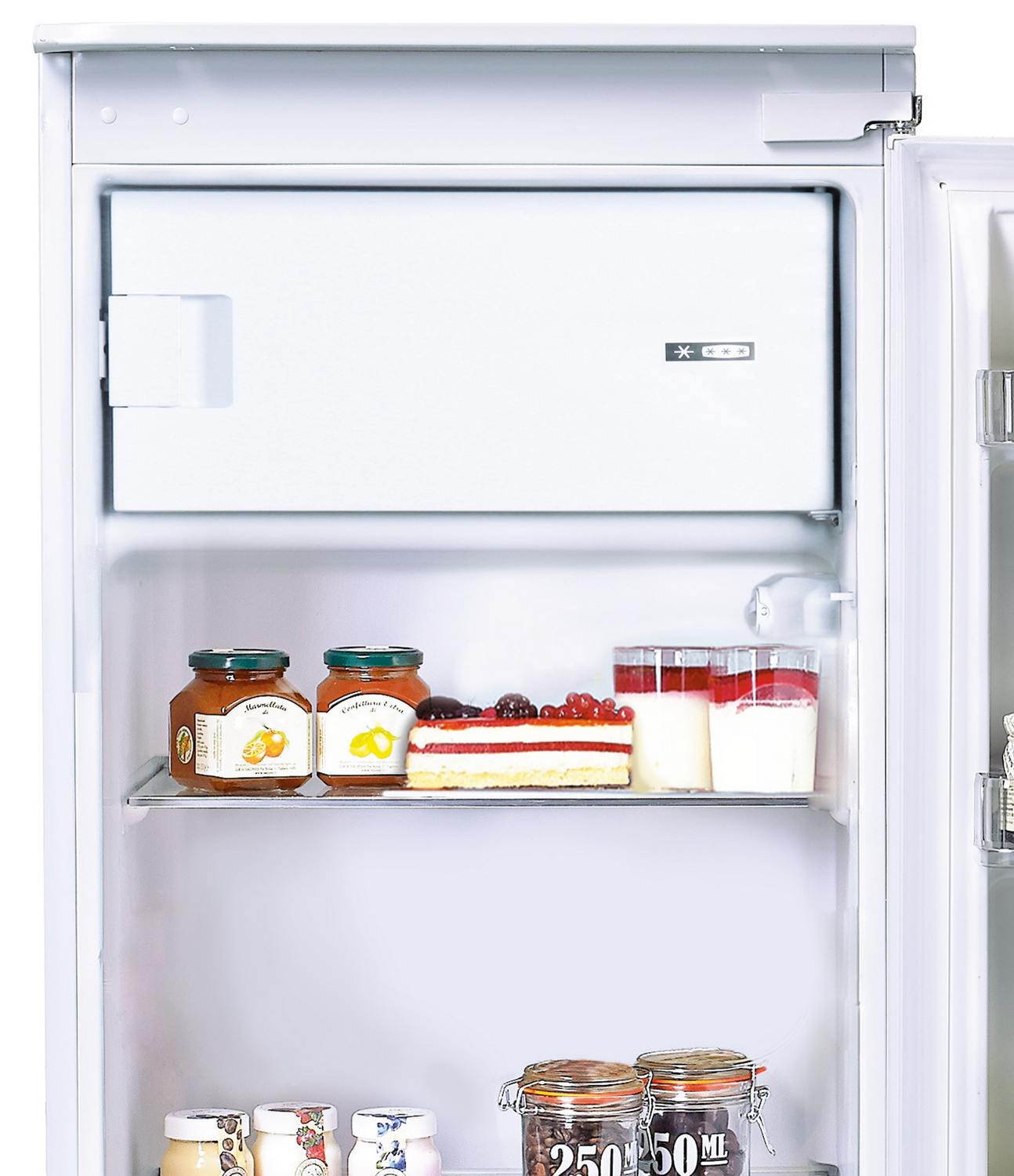 Un refrigerador abierto muestra varios alimentos: frascos de mermelada, pastel, yogur y una sección de congelador. El interior es blanco.