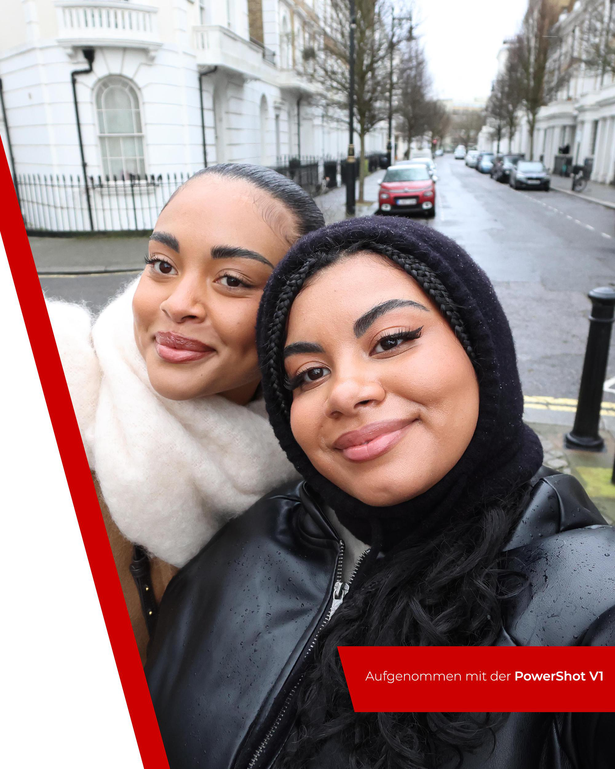 Zwei lächelnde Frauen in einem Selfie. Eine trägt eine schwarze Kapuze und Jacke, die andere einen cremefarbenen Schal. Straße und Autos im Hintergrund.