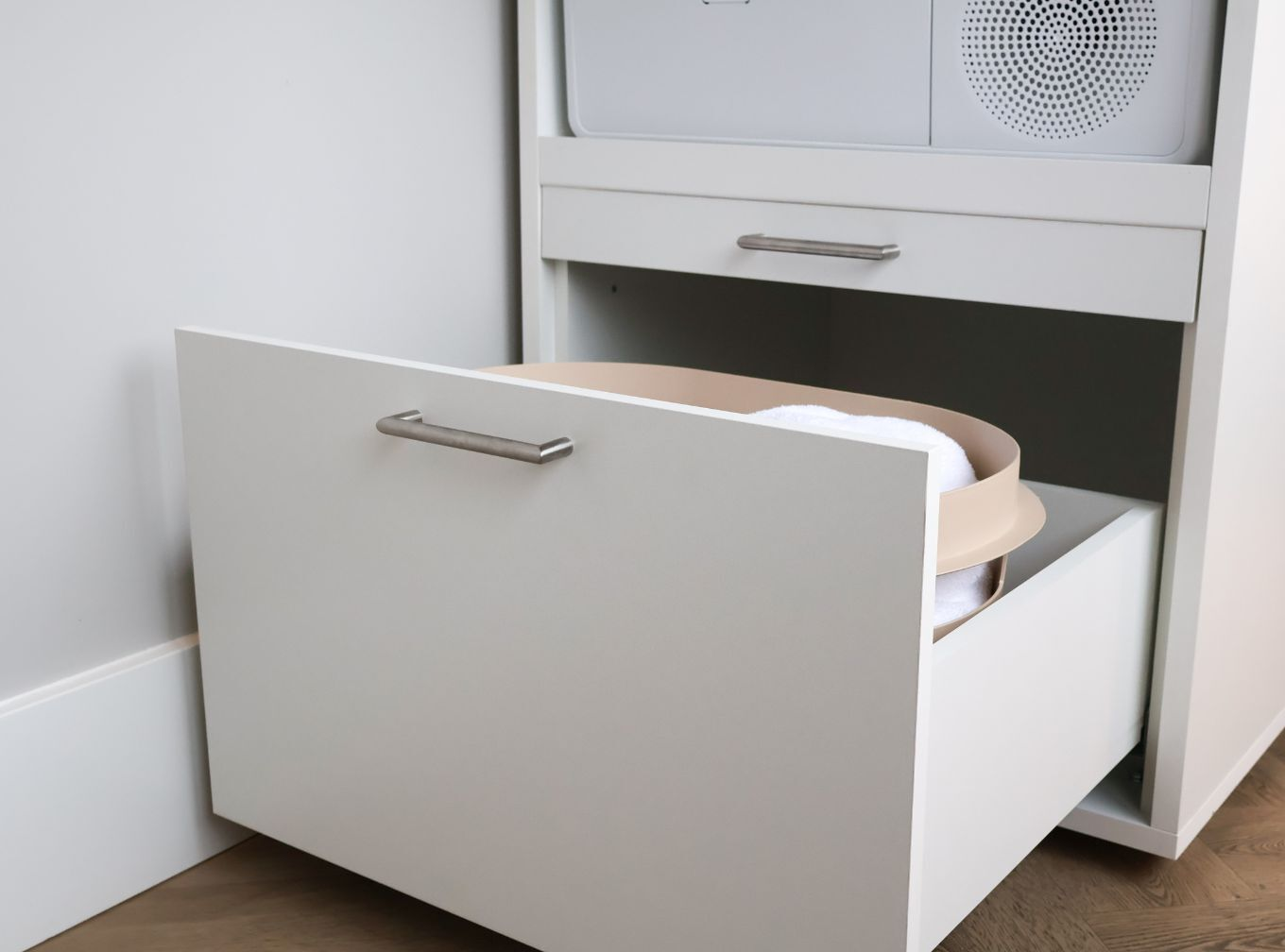 Ein weißer Schrank mit offener Schublade. Darin ein beiger Korb mit weißen Handtüchern. Darüber eine geschlossene Schublade.