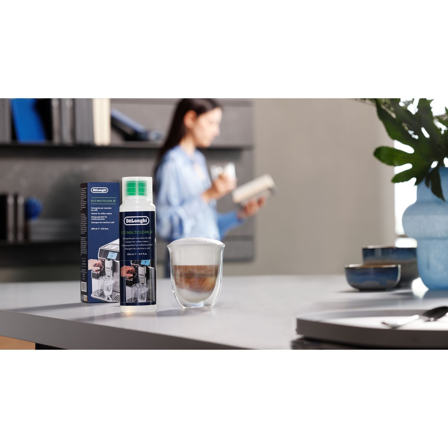 Een DeLonghi-reinigingsfles, een koffiekopje en een vrouw met een boek staan op een aanrecht. Een vaas en kommen staan op de achtergrond.