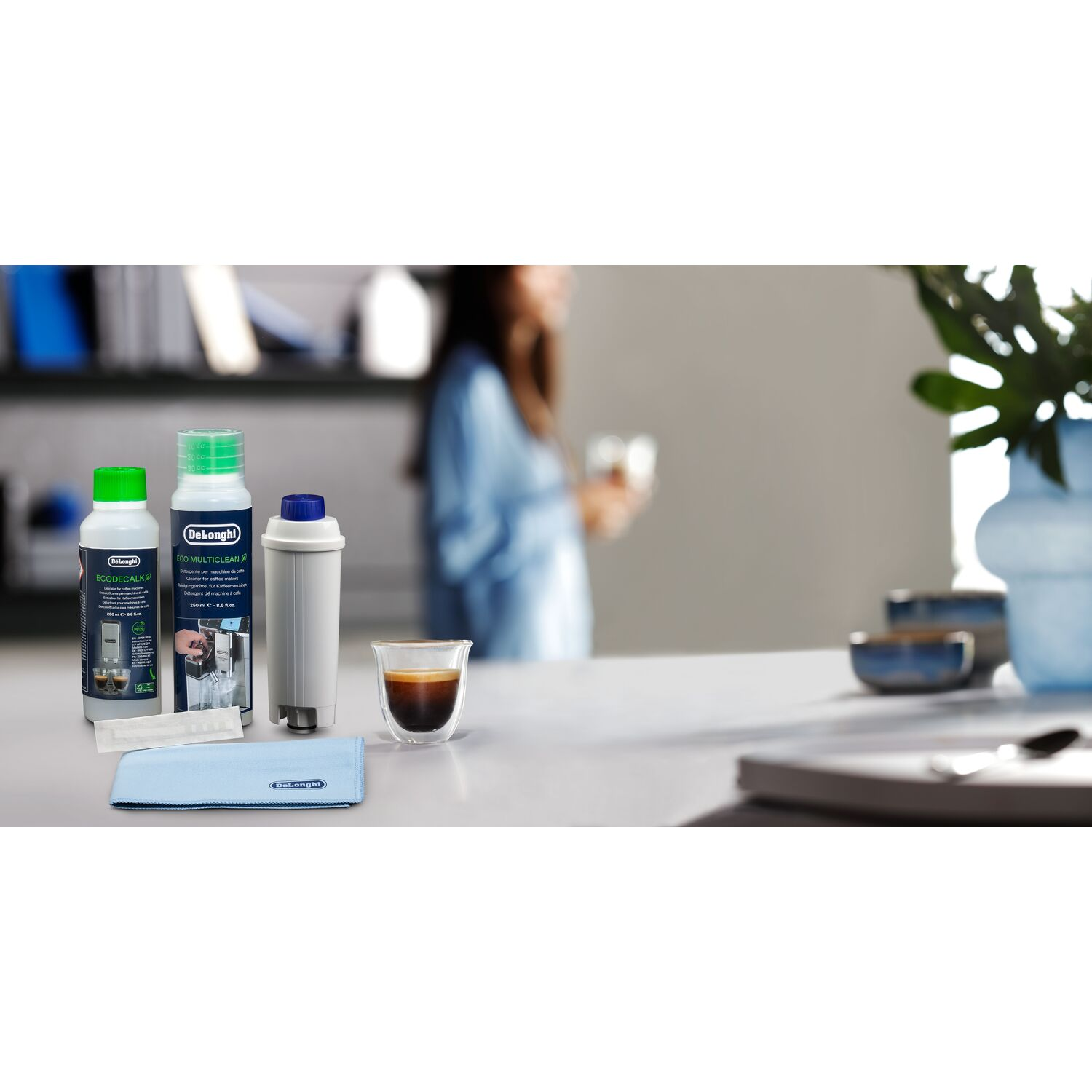 Een tafel met reinigingsproducten voor koffiemachines, een kopje koffie en een vrouw op de achtergrond.
