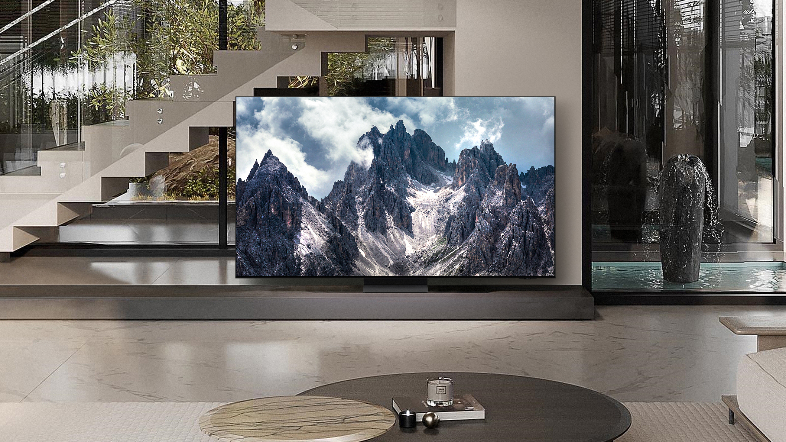 Un moderno soggiorno con TV che mostra montagne. Sono visibili anche una scala, un gioco d'acqua e un tavolino.
