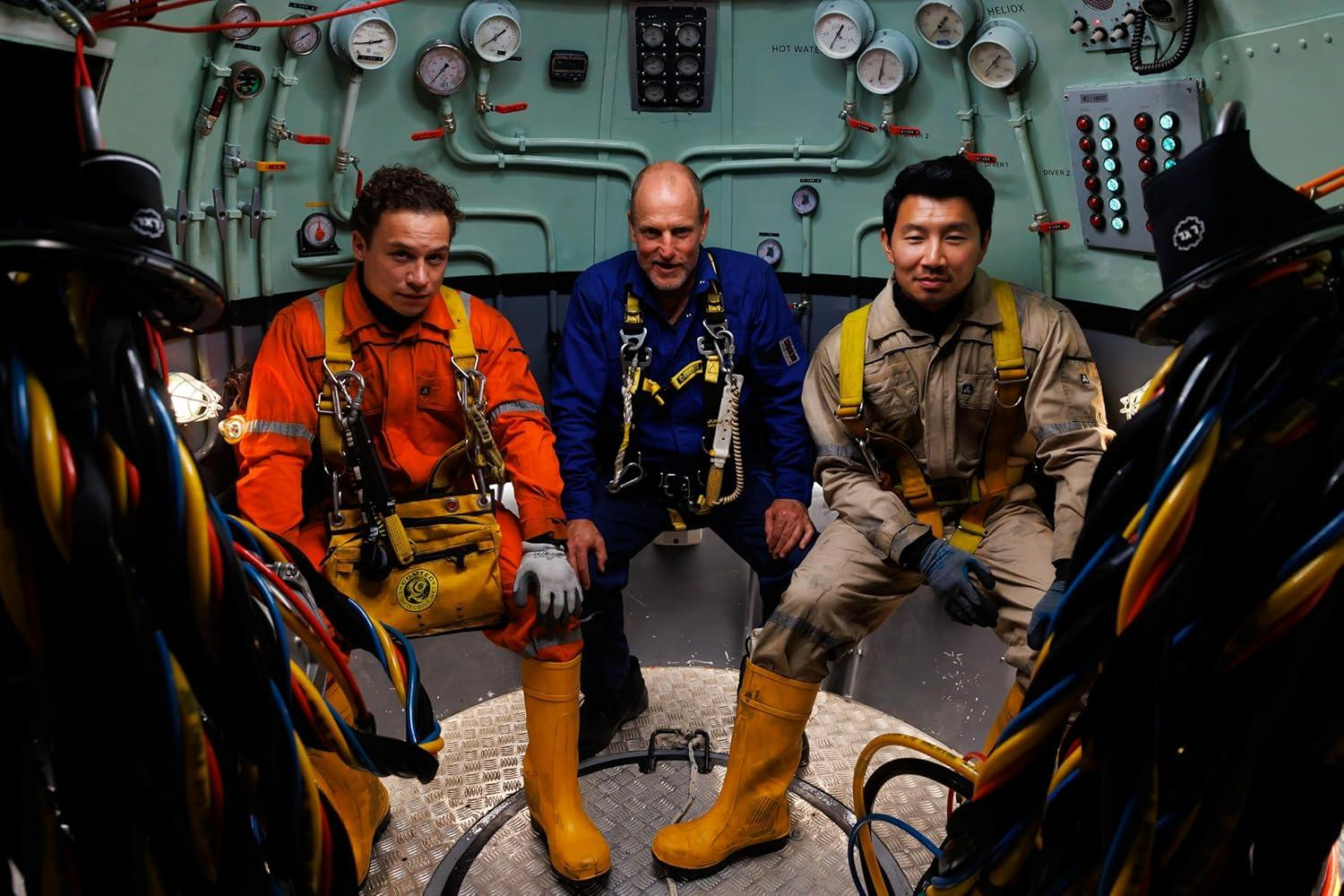 Drei Personen in Arbeitsanzügen sitzen in einem U-Boot. An der Wand befinden sich Anzeigen und Lichter. Kabel sind im Vordergrund.
