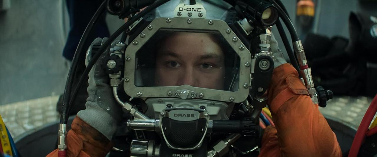 Eine Person mit Taucherhelm, auf dem das Wort "D-ONE" steht. Die Person trägt einen orangenen Anzug.