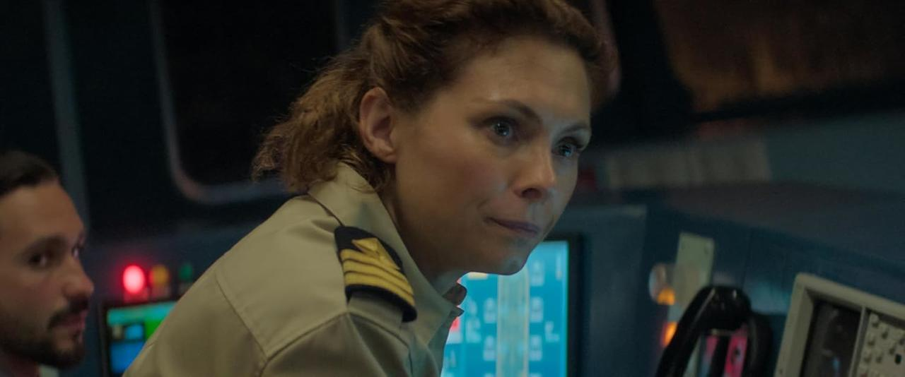 Eine Frau in Uniform blickt zur Seite, links ist ein Mann teilweise zu sehen. Die Szene spielt in einem Kontrollraum.