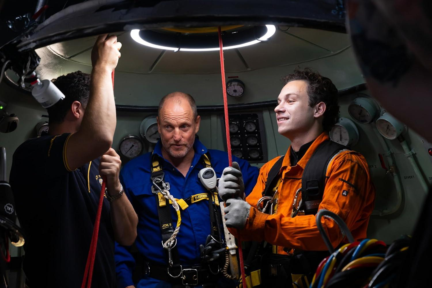 Drei Männer in einem U-Boot. Einer in Orange, einer in Blau. Sie halten rote Seile. Messgeräte und Ausrüstung umgeben sie.