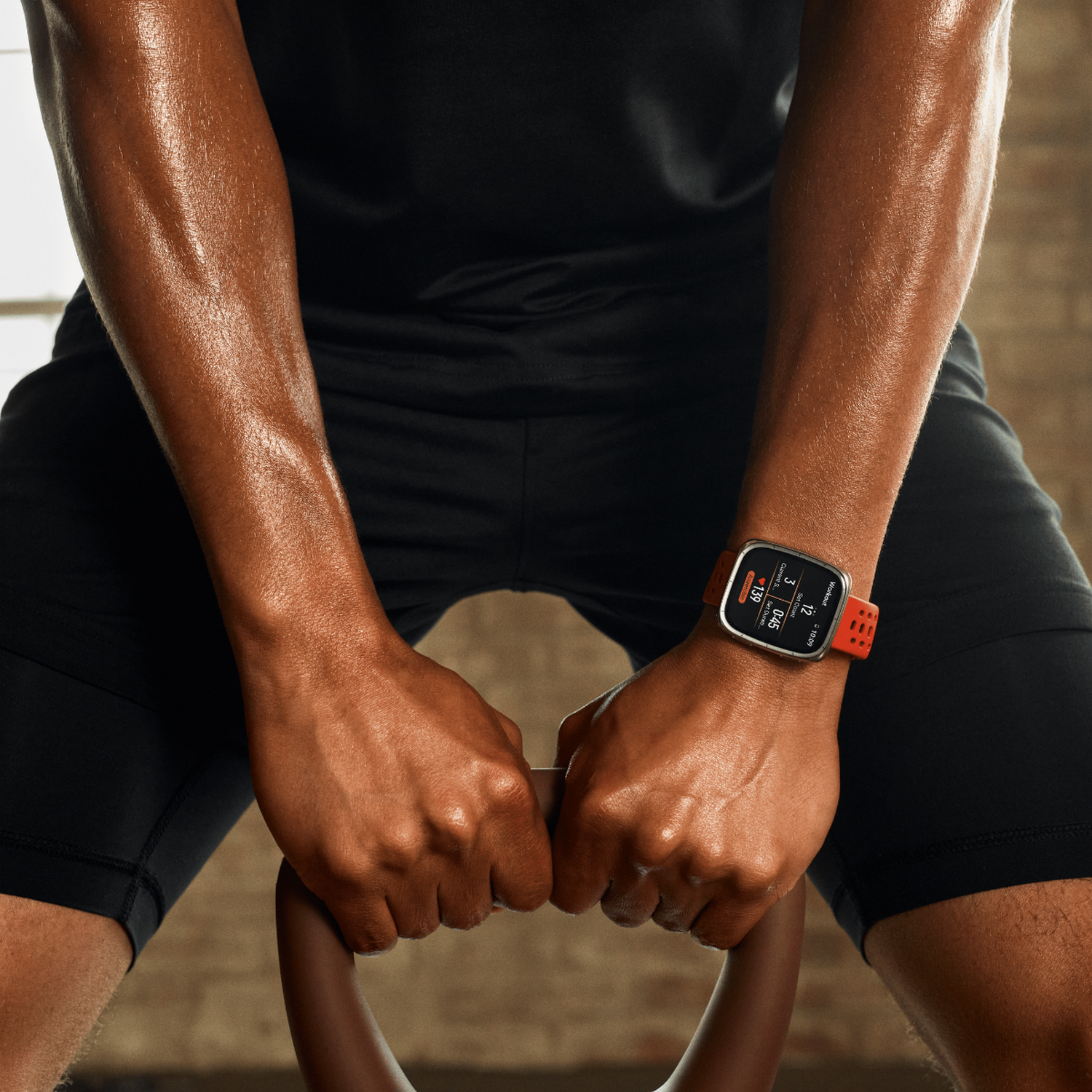 Una persona levanta una pesa rusa. Lleva un reloj inteligente. La persona está sudando y vestida con ropa deportiva negra.