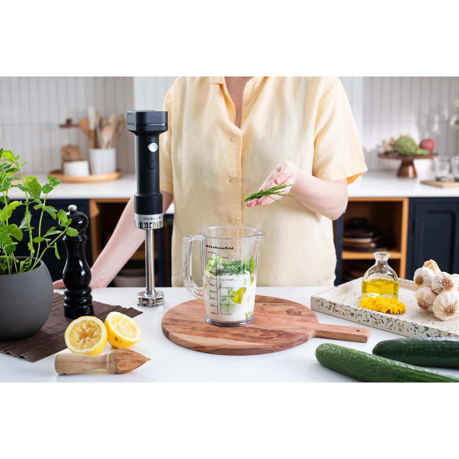 Una persona aggiunge erba cipollina a un frullatore con ingredienti. Limoni, cetrioli, aglio e un frullatore a immersione sul tavolo.