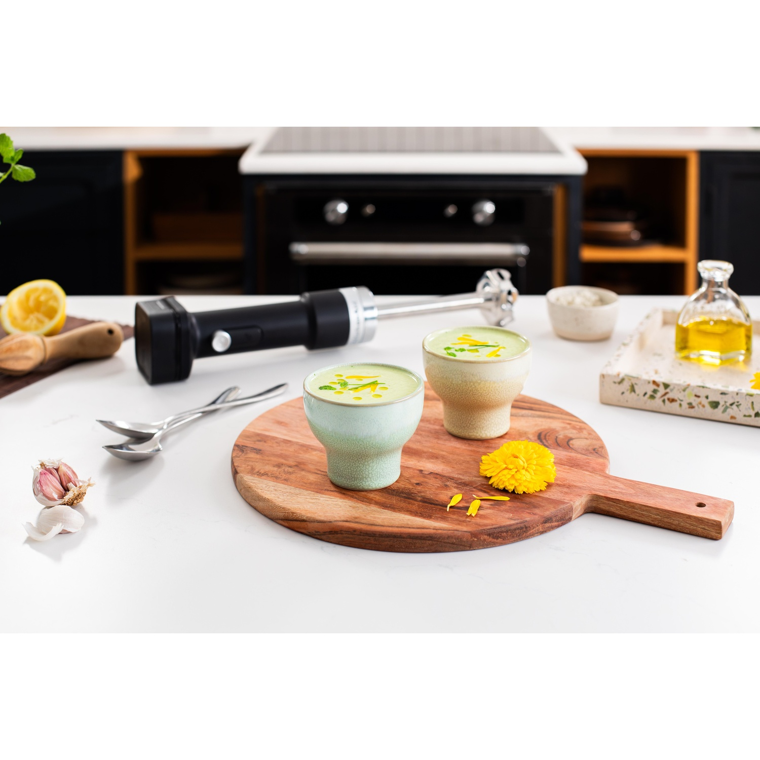 Due ciotole di zuppa su un tagliere di legno. Un frullatore a immersione, aglio, limone e olio sono sul bancone bianco, cucina sullo sfondo.