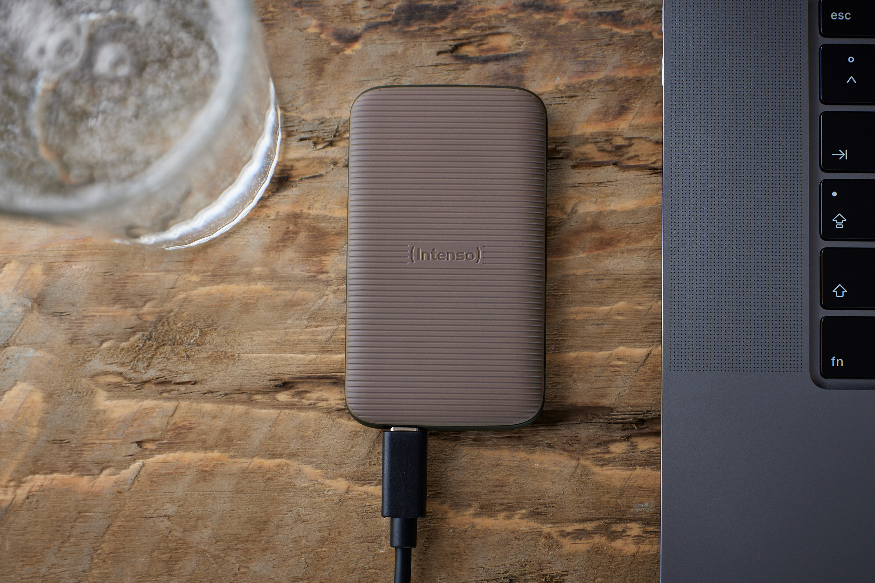 Eine beige externe Festplatte mit schwarzem Kabel ist an einen Laptop auf Holzoberfläche angeschlossen, daneben ein Glas Wasser.