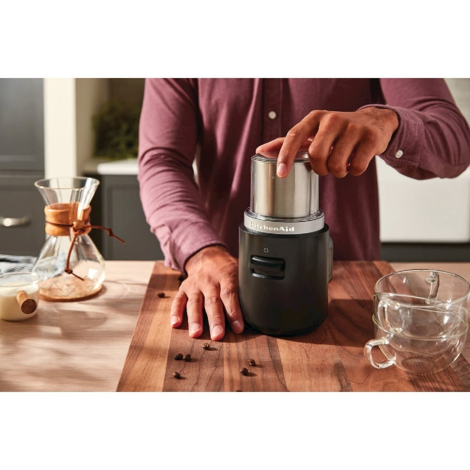 Una persona usa un macinacaffè KitchenAid nero. I chicchi di caffè sono sulla superficie di legno. Sono visibili anche una caraffa e tazze di vetro.