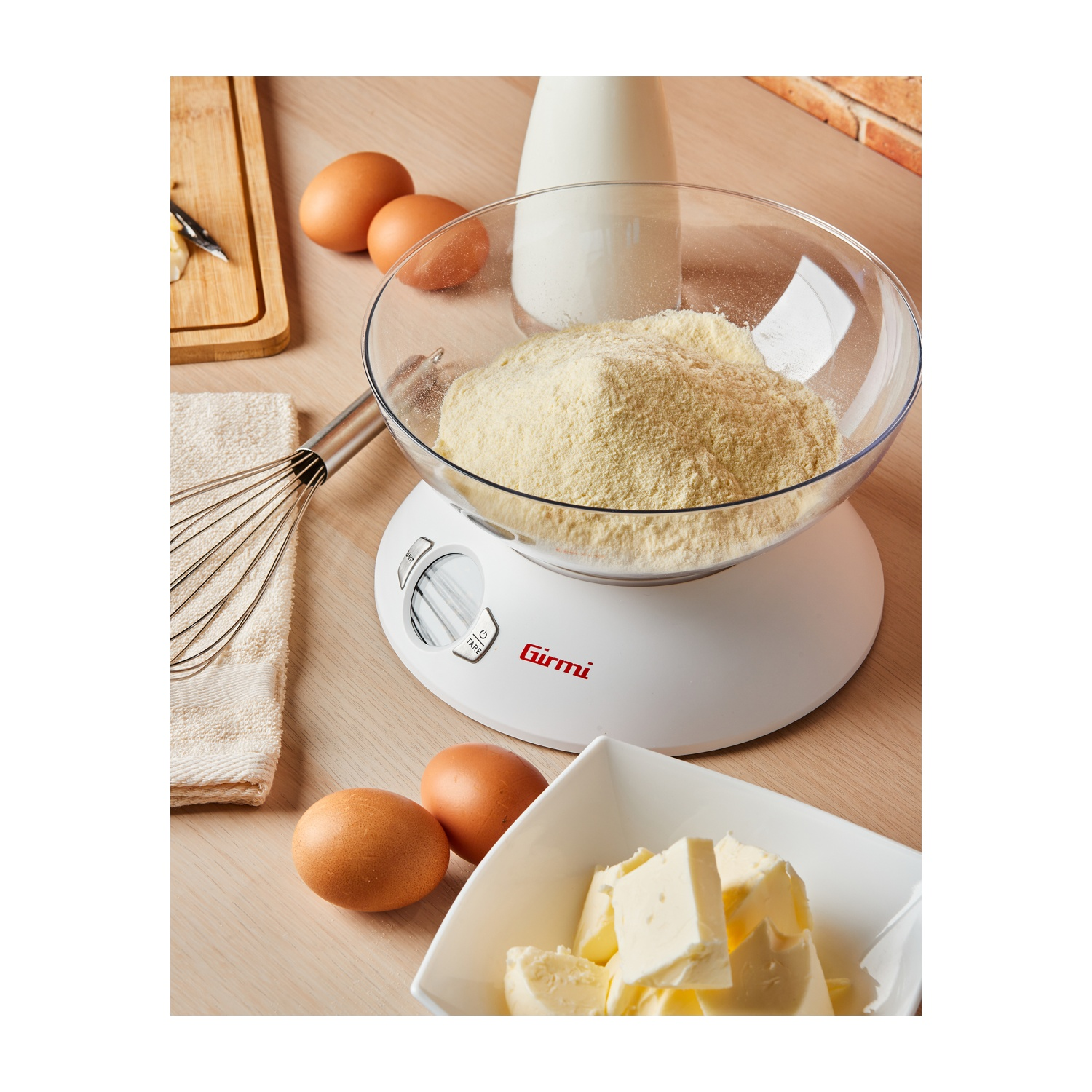 Una scena in cucina con una bilancia che contiene farina. Uova, burro, latte e una frusta sono disposti su una superficie di legno.