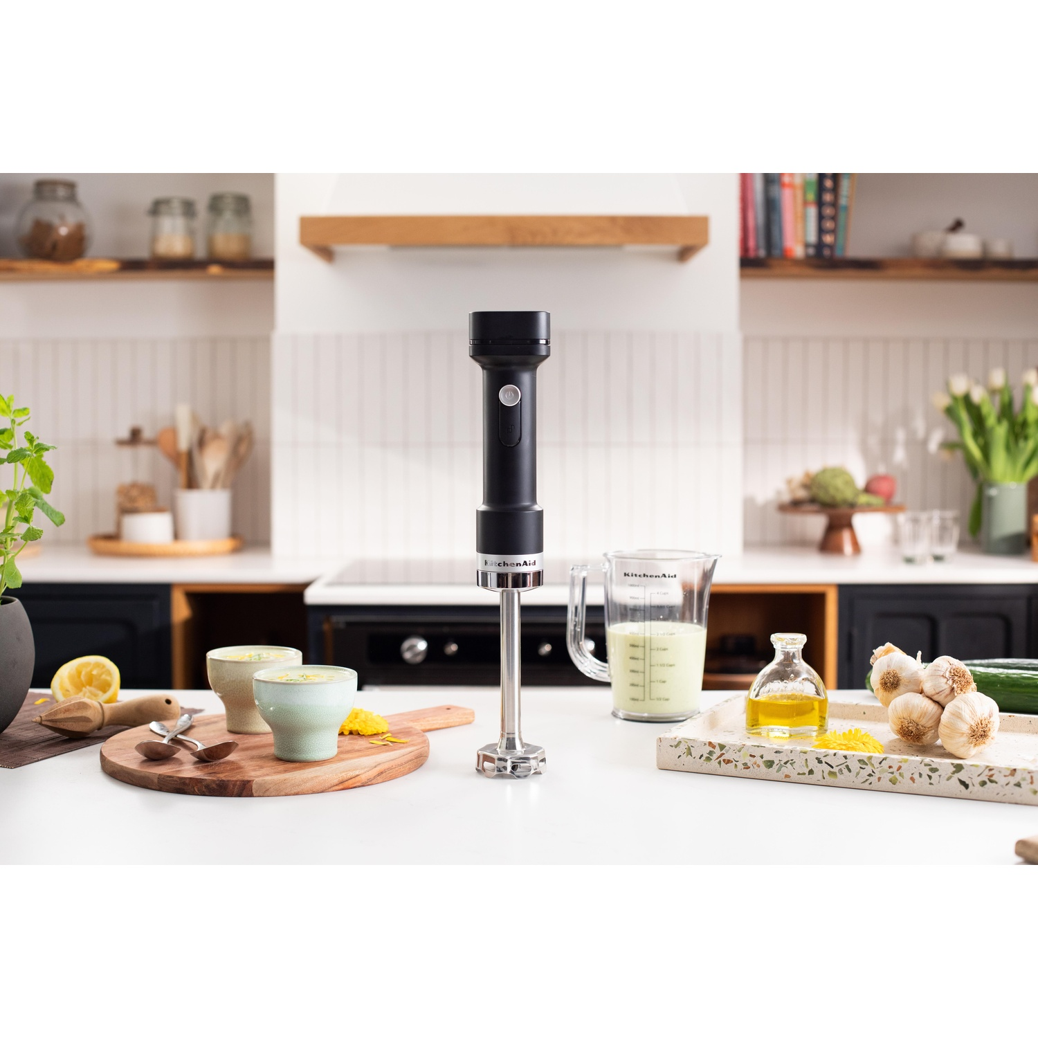 Una scena di cucina con un frullatore a immersione nero. Tazze di zuppa, aglio, olio e un misurino sono sul bancone.