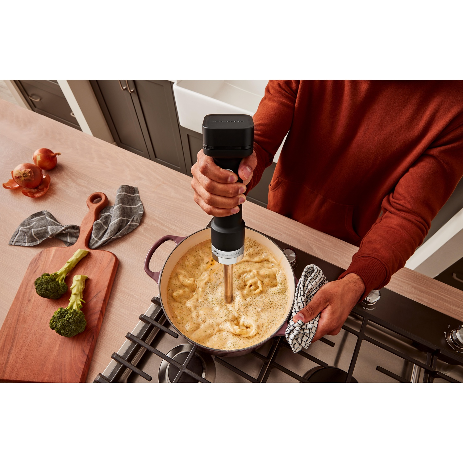 Una persona frulla la zuppa in una pentola viola con un frullatore a immersione. Broccoli, cipolle e un tagliere in legno sono vicini.