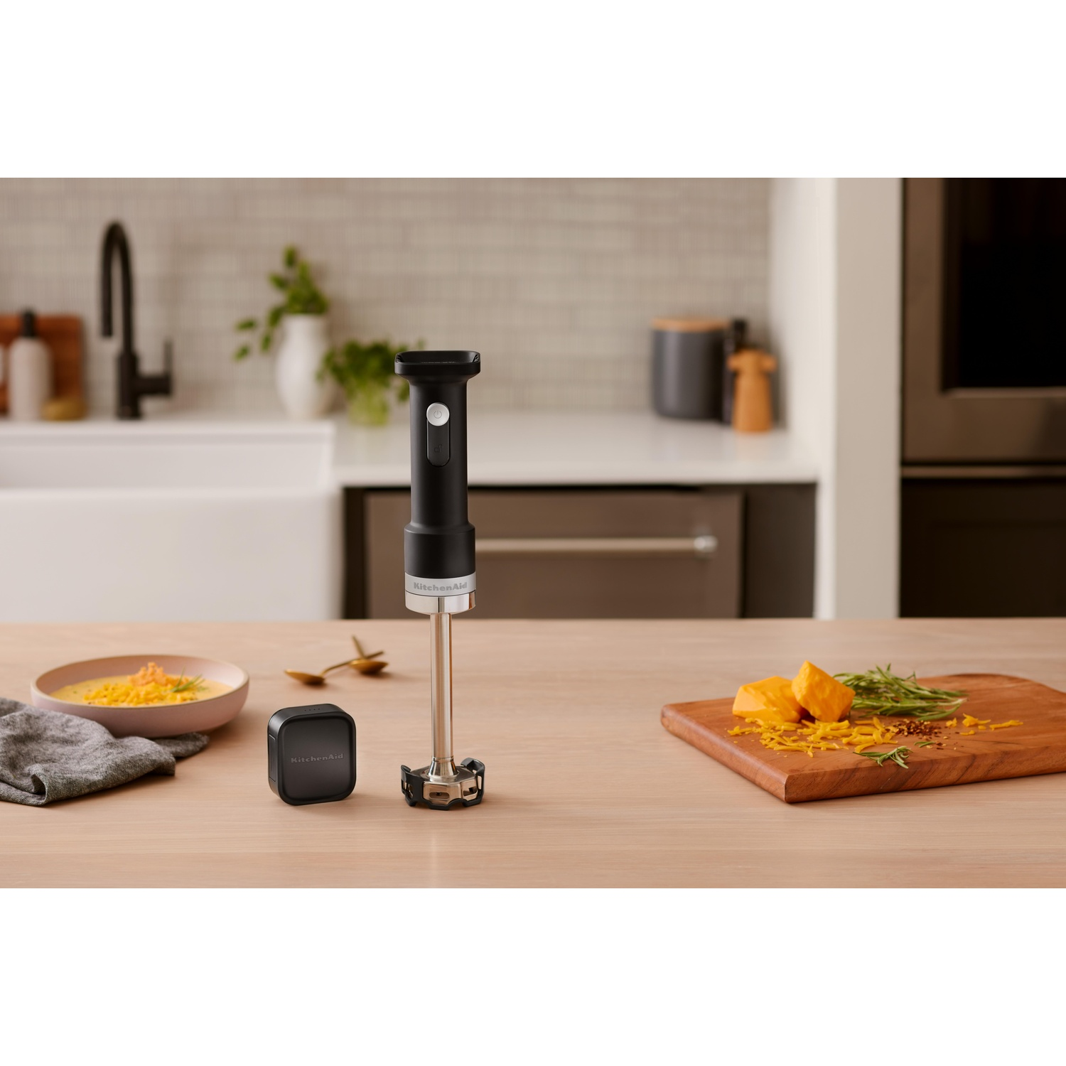 Un frullatore a immersione KitchenAid nero è posizionato su un tavolo di legno. Sono presenti anche zuppa, formaggio e rosmarino, con un tagliere di legno.