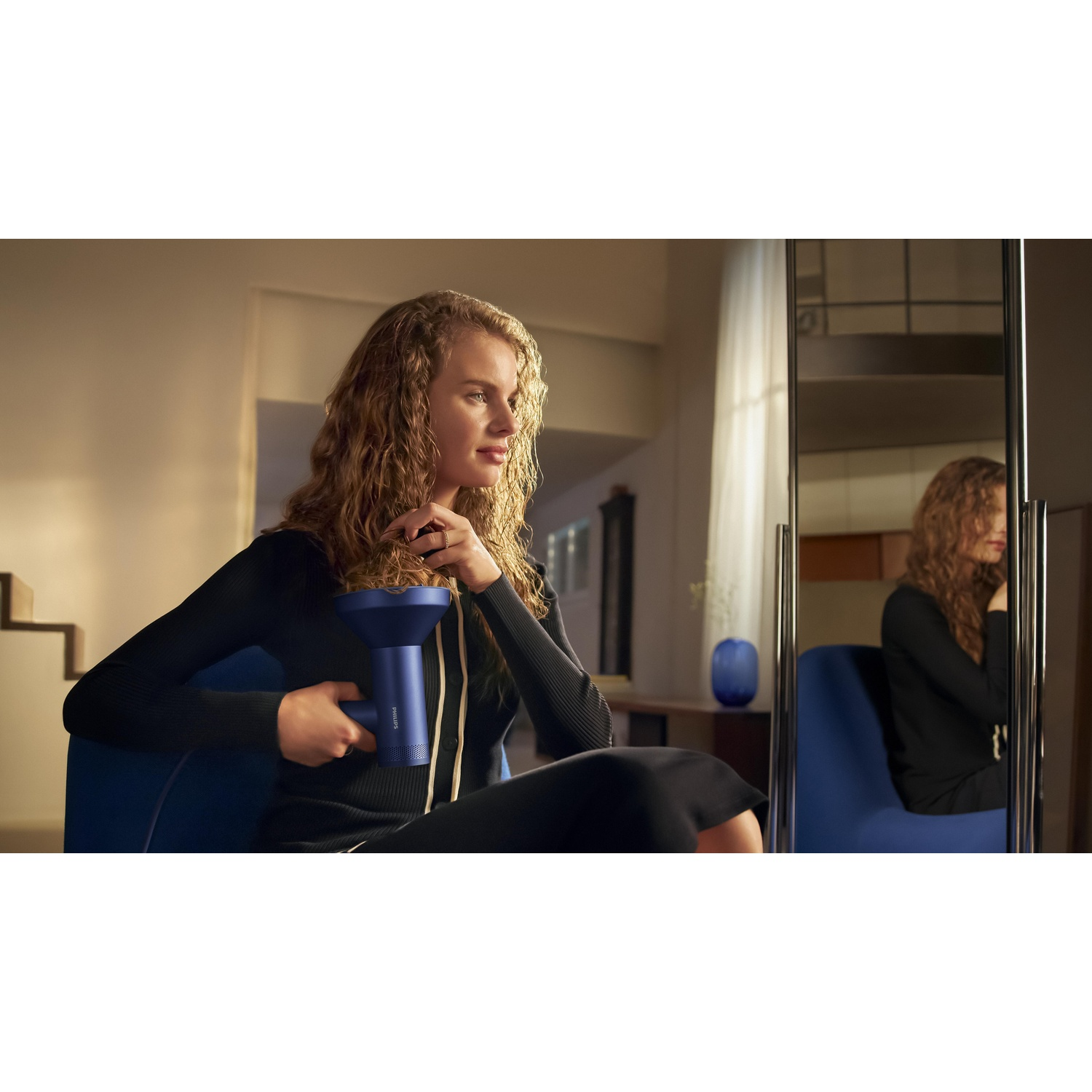 Una donna con i capelli ricci tiene un asciugacapelli blu, seduta su una sedia blu. Uno specchio la riflette. Indossa un vestito nero.