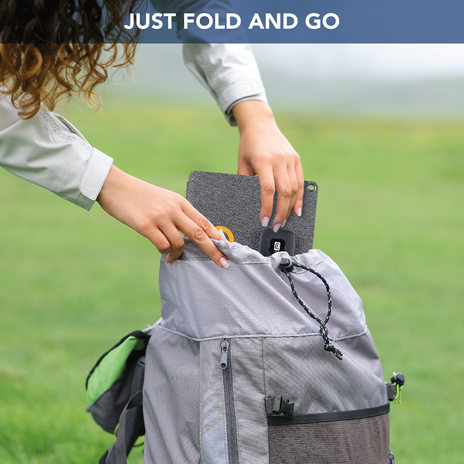 Una persona coloca un artículo gris doblado en una mochila gris. El fondo es un campo verde, con el texto "JUST FOLD AND GO" encima.