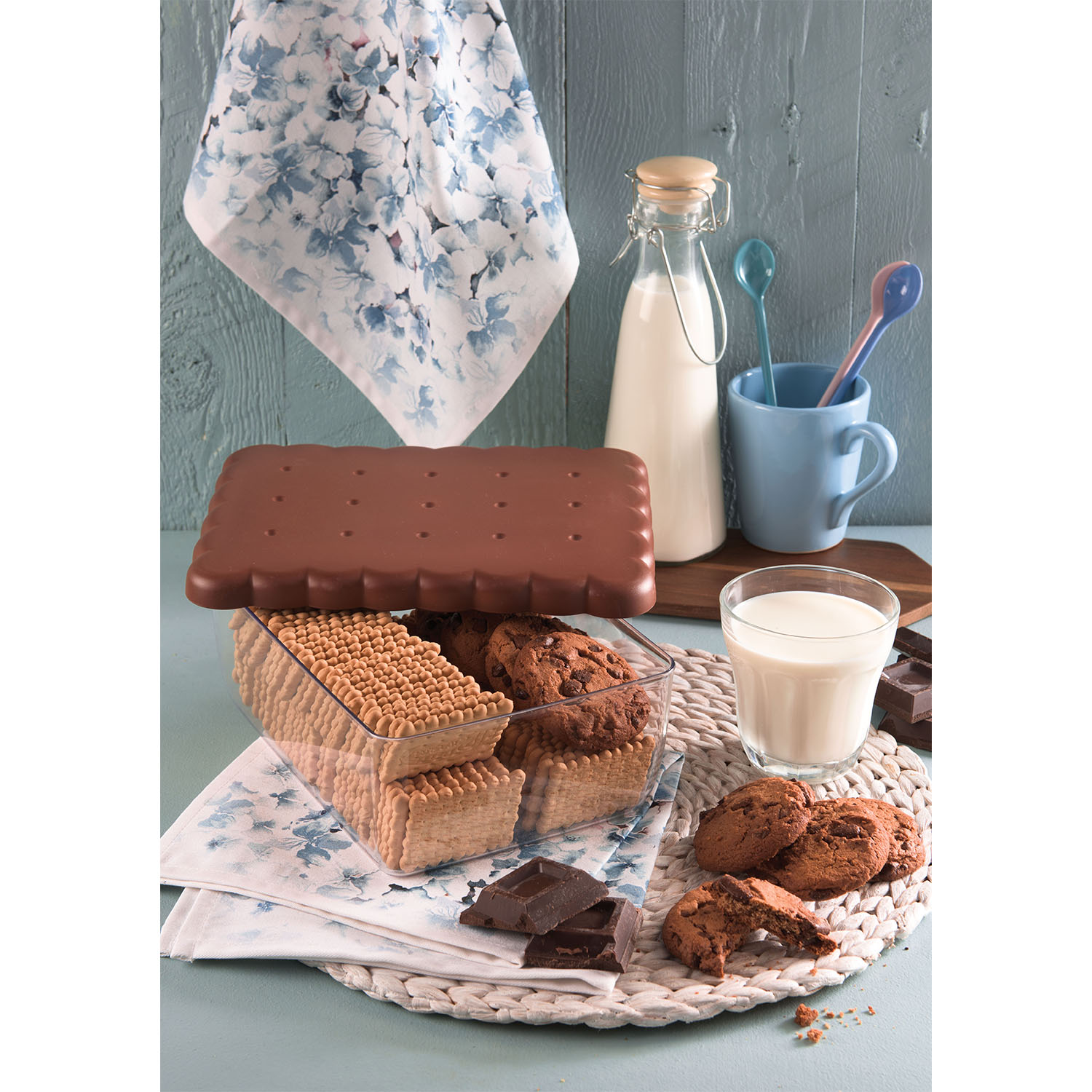 Biscotto al cioccolato, pasticcini, latte in un bicchiere, bottiglia e tazza, su una superficie di legno con un tovagliolo floreale.