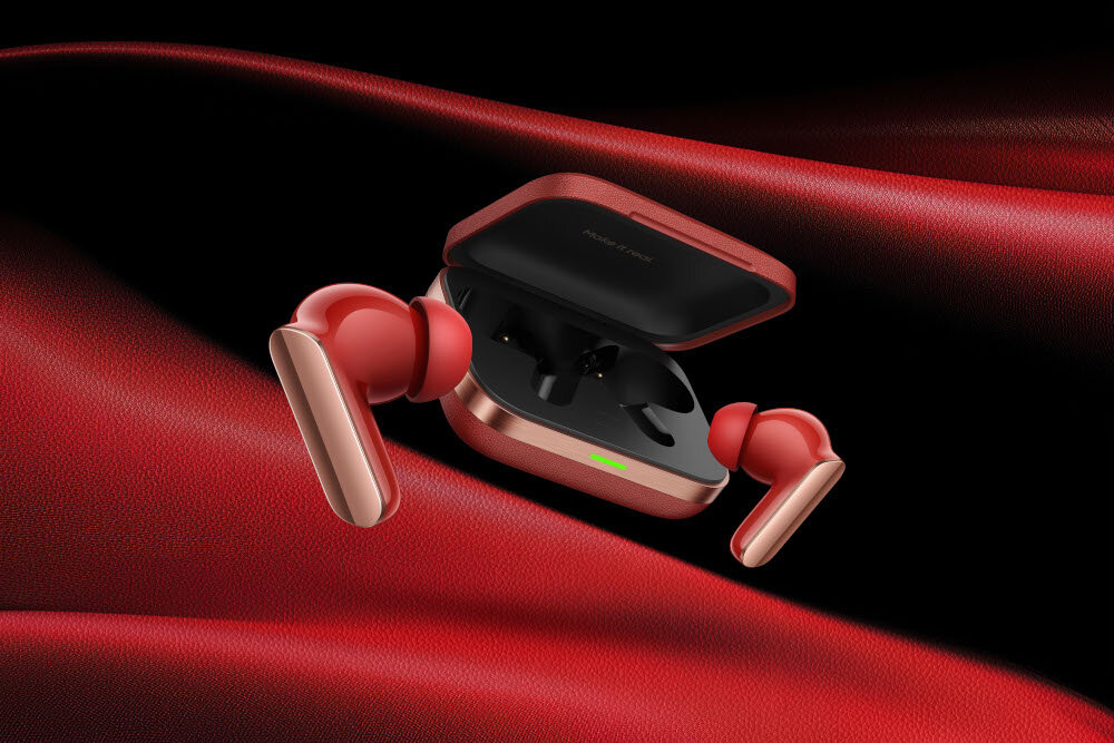 Czerwone i złote bezprzewodowe słuchawki, etui otwarte. Słuchawki znajdują się poza etui. Czerwone tło z tkaniny z czarnym w prawym górnym rogu.