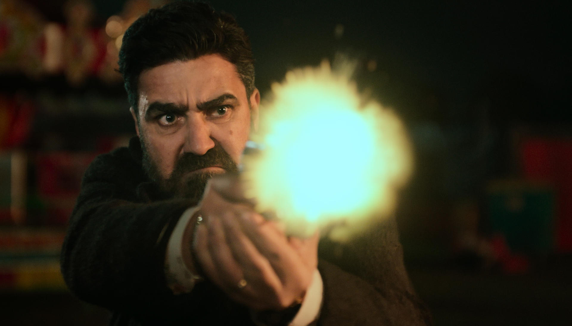 Ein Mann mit Bart schießt mit einer Pistole. Heller Lichtblitz geht von der Waffe aus. Dunkler Hintergrund. Der Mann ist konzentriert und intensiv.