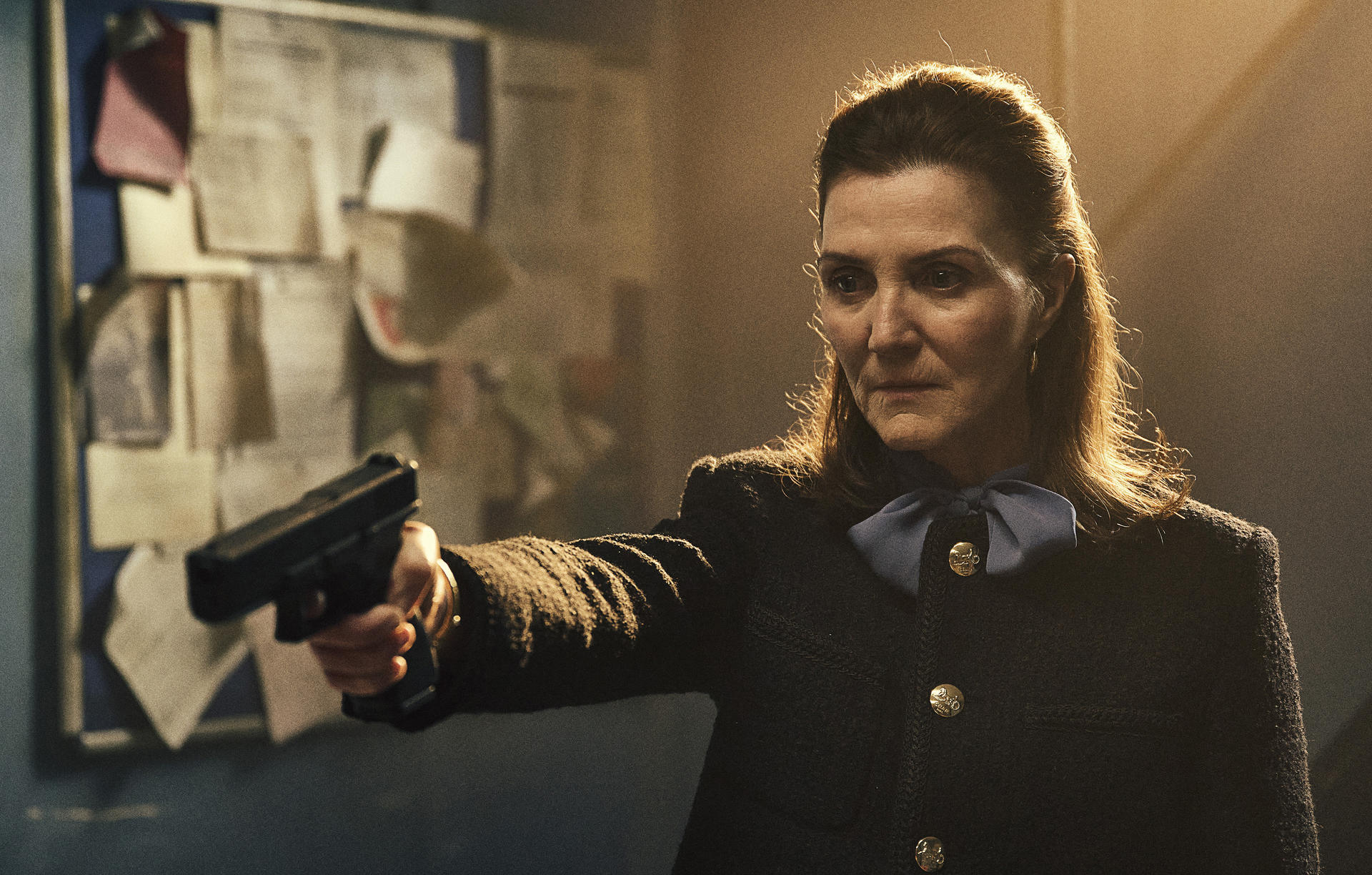 Eine Frau in einem Blazer hält eine Waffe. Sie hat eine blaue Schleife und steht in der Nähe einer Pinnwand. Der Hintergrund ist gedämpft.
