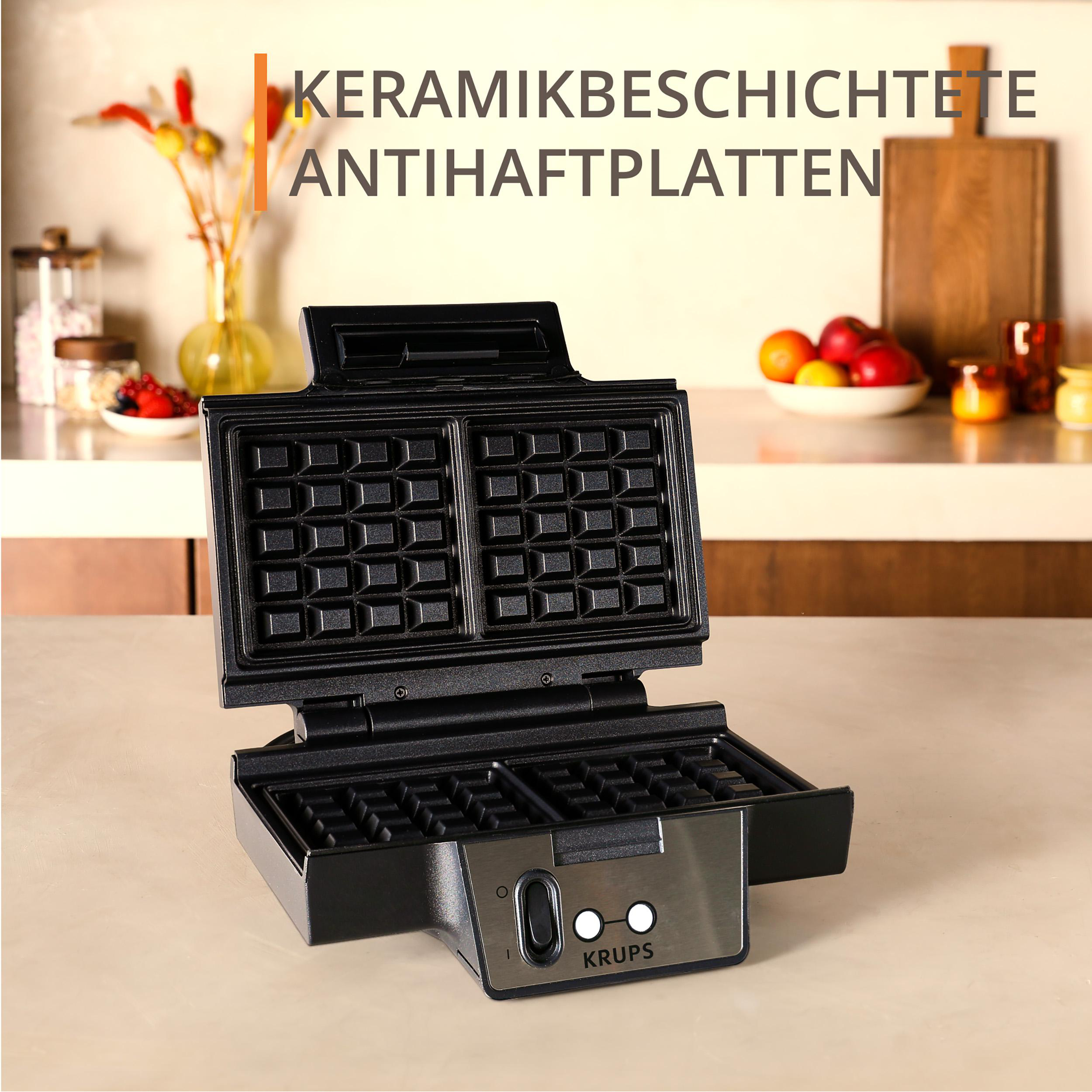 Ein schwarzer Waffeleisen steht auf einer Theke. Dahinter sind Früchte und Blumen. Auf dem Waffeleisen steht das Wort KRUPS.