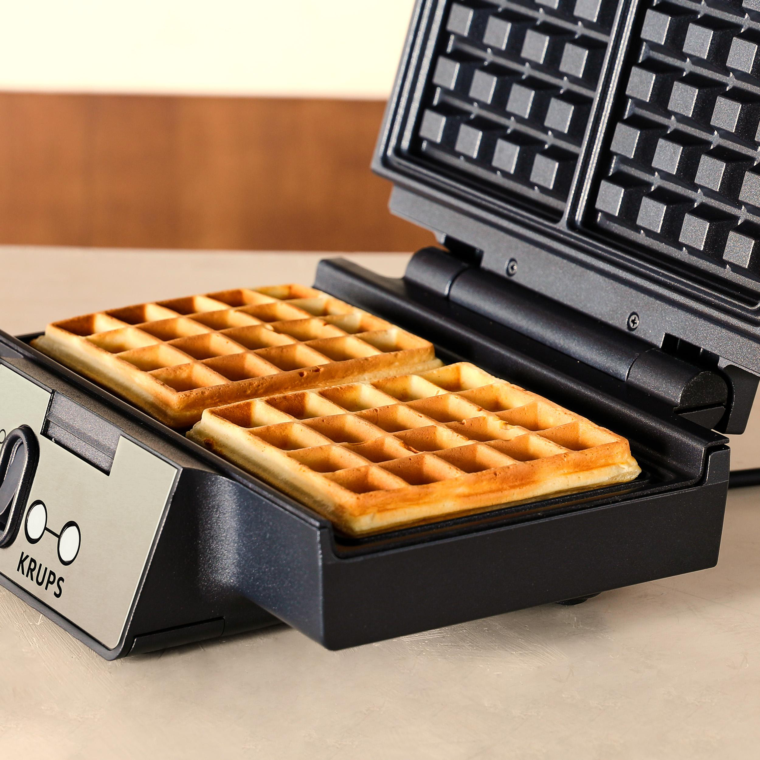 Waffeleisen mit zwei goldenen Waffeln. Das Gerät ist geöffnet und zeigt die gekochten Waffeln. Der Hintergrund ist unscharf.