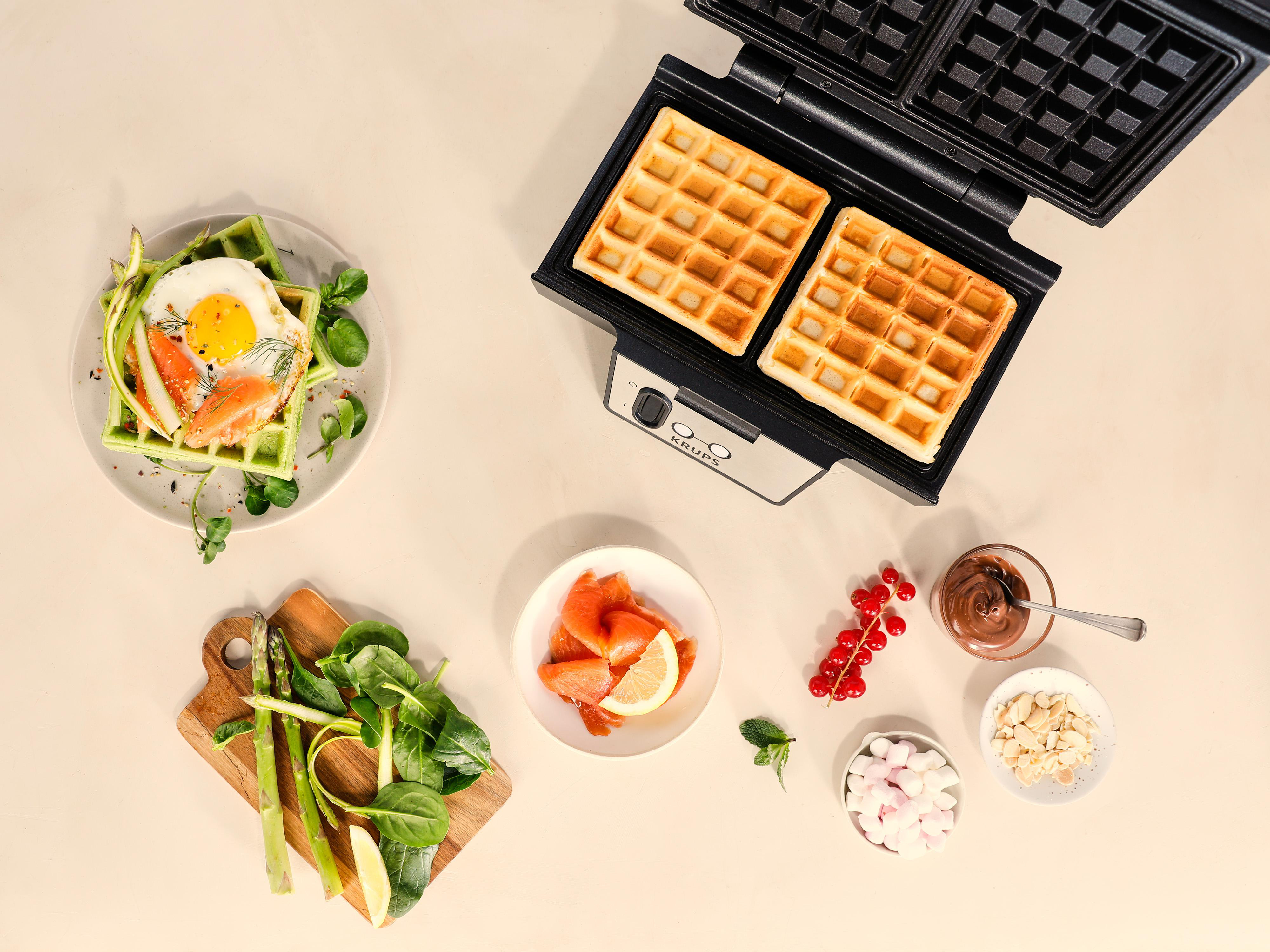 Draufsicht zeigt Waffeln, Zutaten und einen Waffeleisen. Waffeln, Ei, Räucherlachs, Beeren und Nüsse sind sichtbar.
