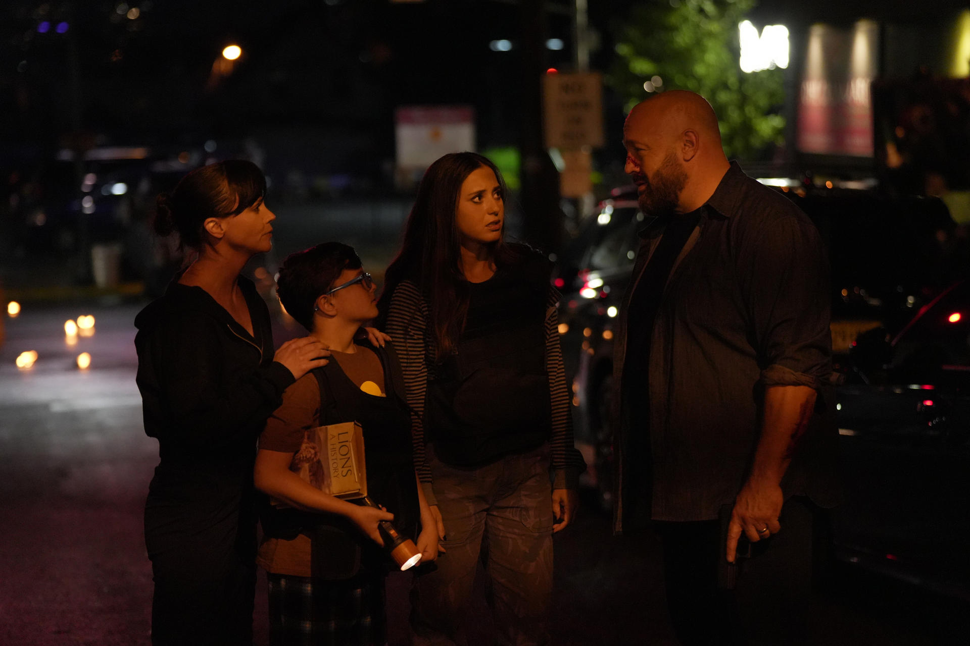 Eine Familie steht nachts auf der Straße. Ein Mann mit Bart redet mit ihnen. Einer hält eine Taschenlampe und ein Buch.