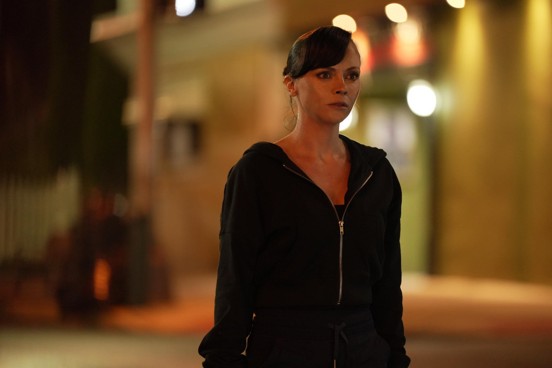 Eine Frau in einem schwarzen Kapuzenpullover steht nachts draußen. Der Hintergrund ist mit Straßenlaternen und Gebäuden verschwommen.