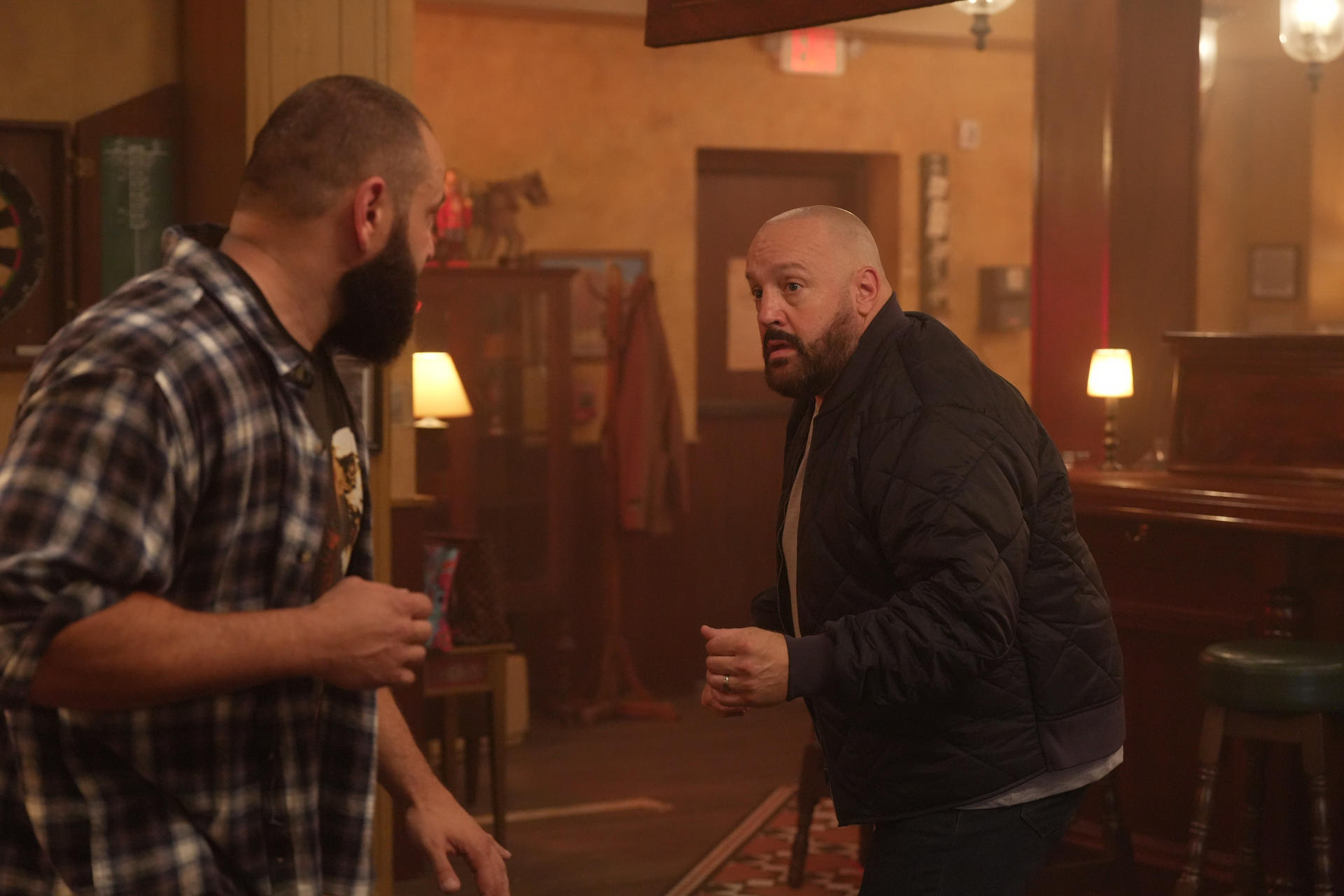 Zwei Männer in einer Bar. Einer trägt ein kariertes Hemd, der andere eine schwarze Jacke. Sie unterhalten sich. Hintergrund Bar-Dekor.