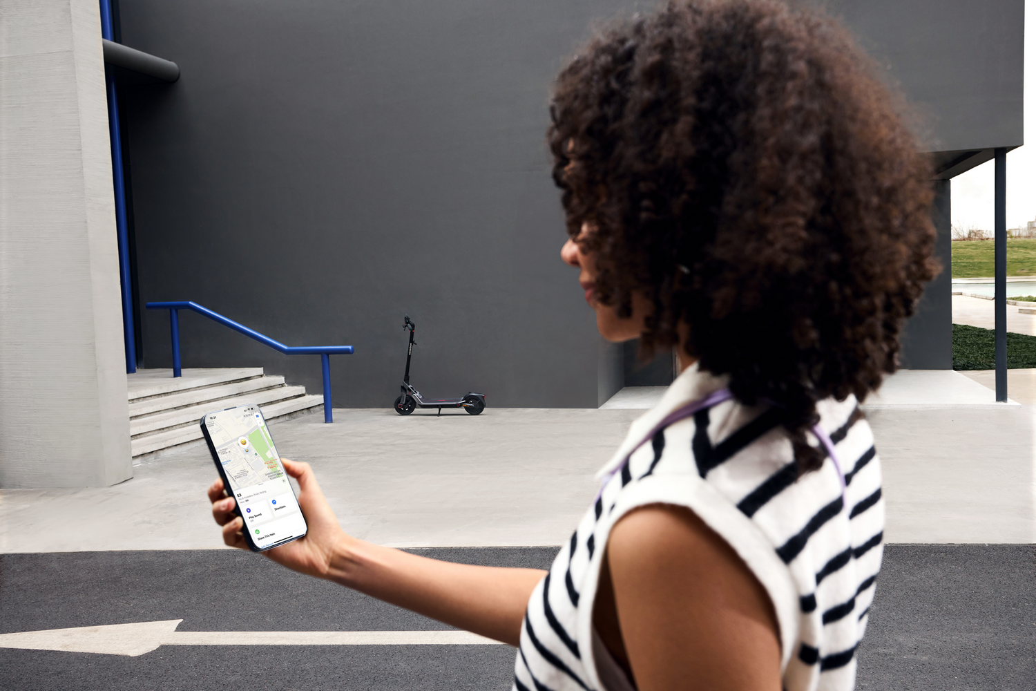 Mujer usando teléfono con mapa, cerca de un scooter. Fondo: pared gris, escaleras, barandilla azul y zona verde.