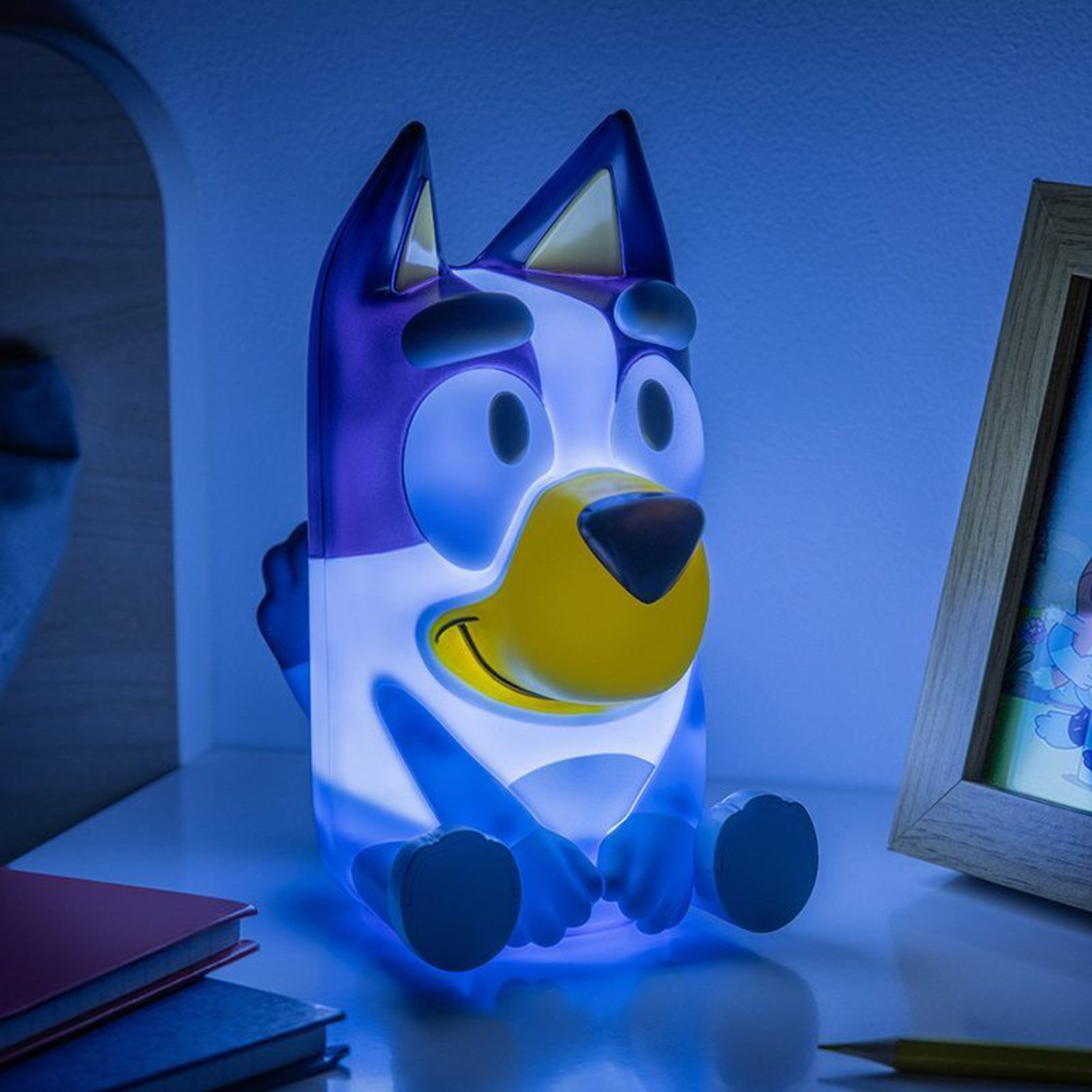 A Bluey night light sits on a shelf. The light is blue and white. A picture frame and books are also on the shelf.