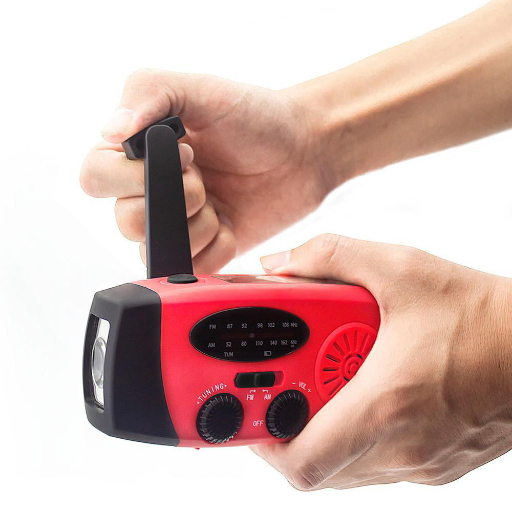 Eine Person hält ein rotes Handkurbelradio mit schwarzem Griff. Es hat eine Taschenlampe und Drehregler. Der Hintergrund ist weiß.