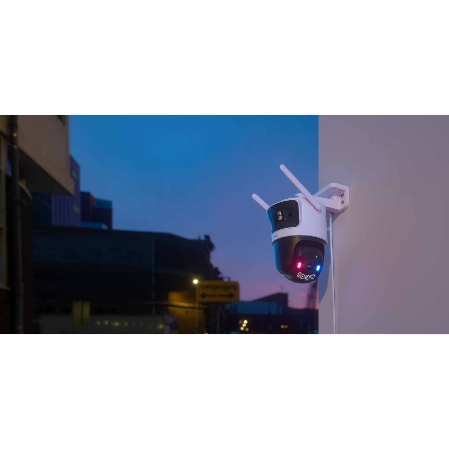 A white security camera with antennas is mounted on a wall. The background shows a street at dusk with buildings and a blue sky.