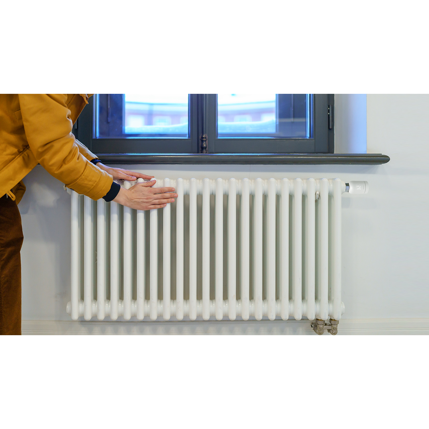 A person in a yellow jacket touches a white radiator below a window. The radiator is against a white wall.