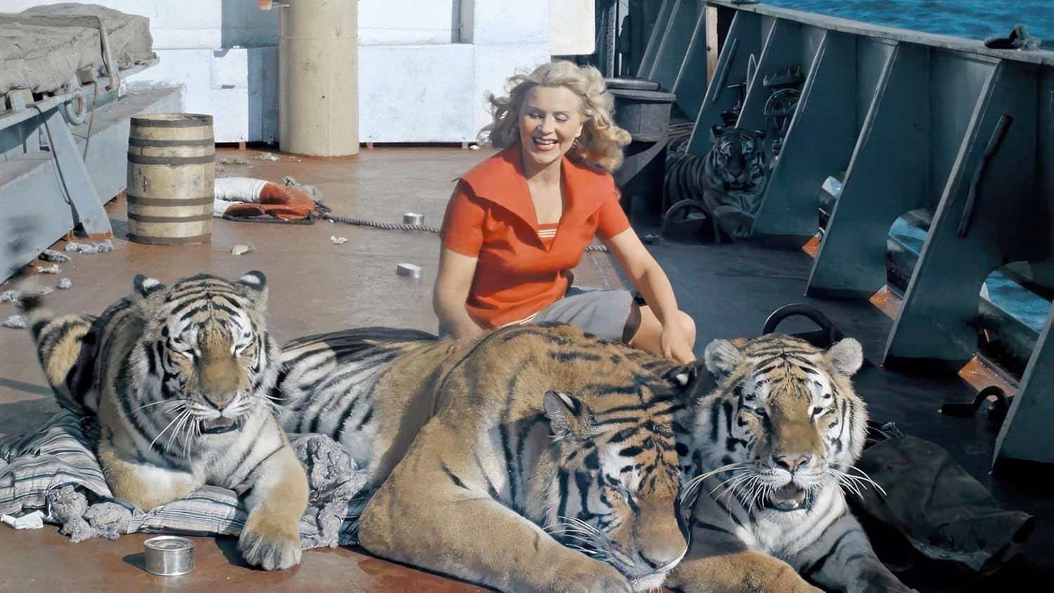 Eine Frau in Rot sitzt mit drei Tigern auf einem Bootsdeck. Ein vierter Tiger ist im Hintergrund. Der Himmel und das Meer sind sichtbar.