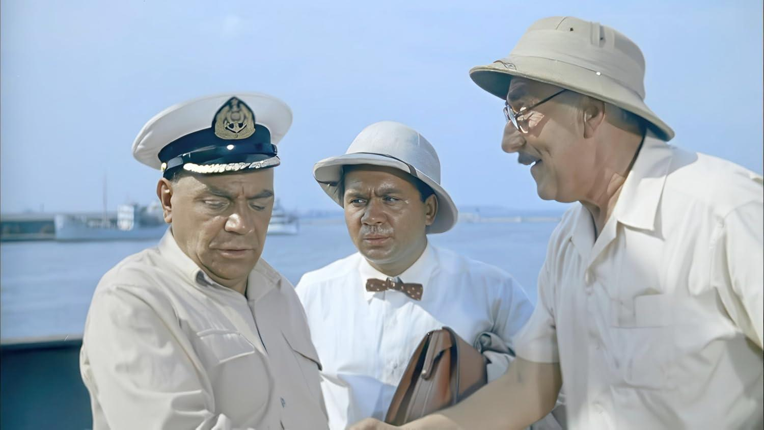 Drei Männer mit Hüten und hellen Hemden stehen auf einem Schiffsdeck. Im Hintergrund sind Wasser und entfernte Gebäude zu sehen.