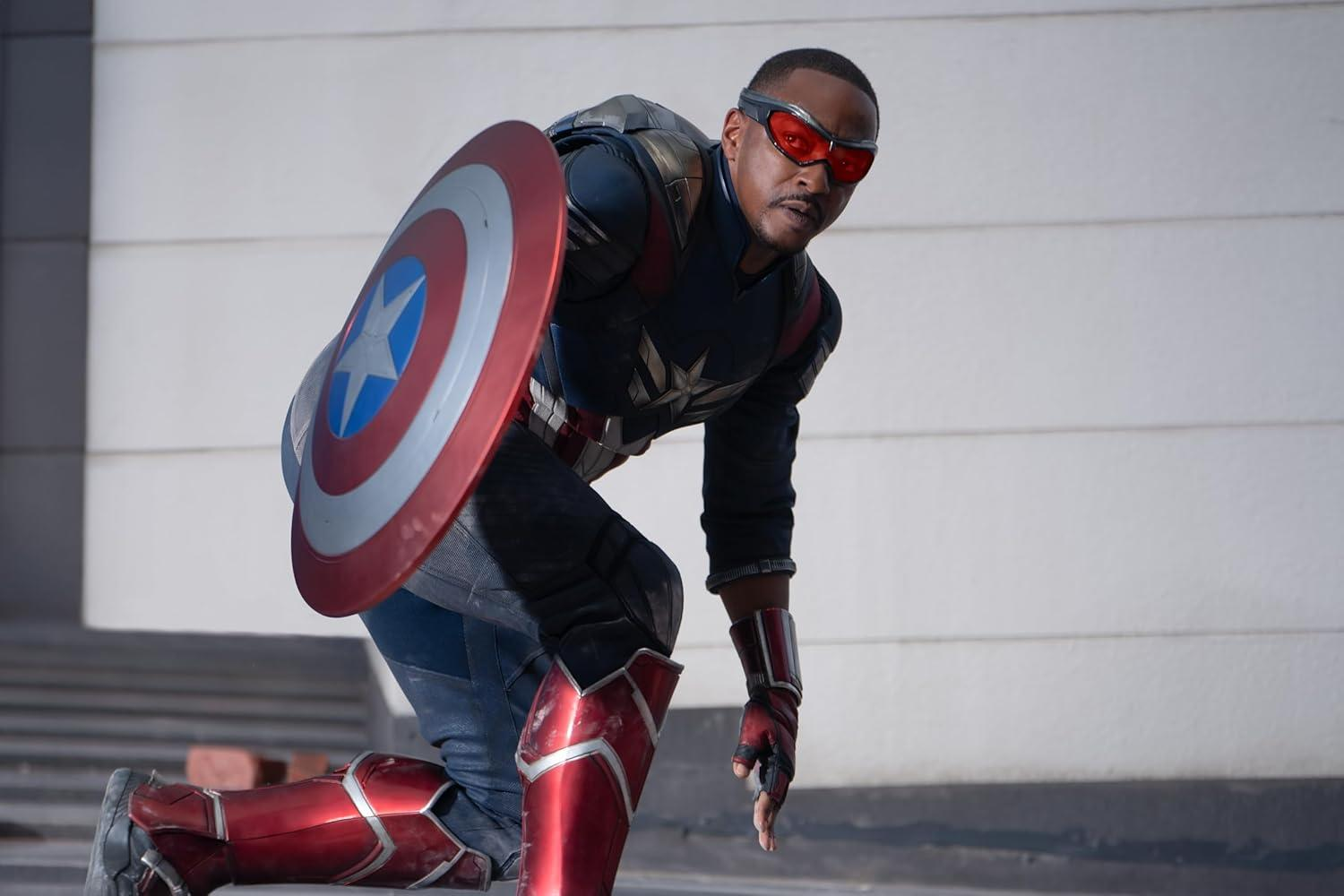 Mann im Captain America-Anzug kniet, Schild haltend. Rote Schutzbrille und Stiefel. Blau-roter Anzug. Weißer Wandhintergrund.