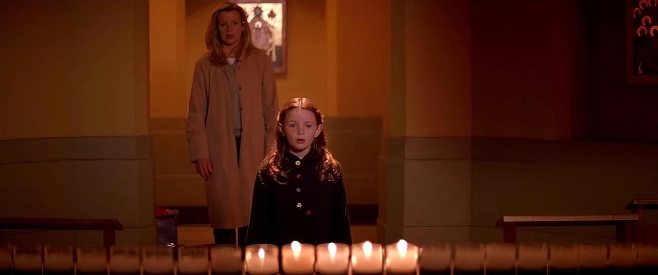 Eine Frau und ein Mädchen stehen in einer Kirche. Das Mädchen steht vor Kerzen. Die Frau trägt einen beigen Mantel.