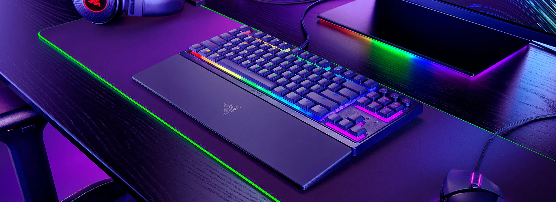 Egy fekete gamer billentyűzet színes fényekkel, egérrel és fejhallgatóval az asztalon. A Razer logó látható.